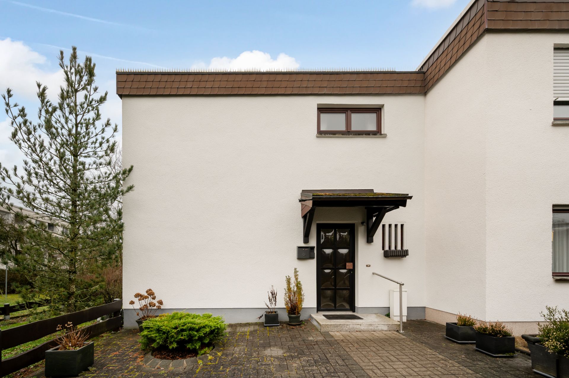 Weiße Hausfassade mit brauner Tür, kleinem Fenster oben und bepflanzten Kübeln am Eingang.
