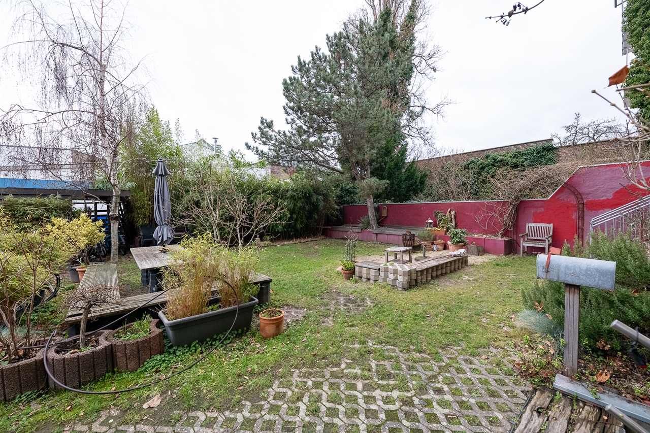 Garten mit Sitzbereich, Bäumen, Pflanzenkübeln und roter Mauer im Hintergrund.