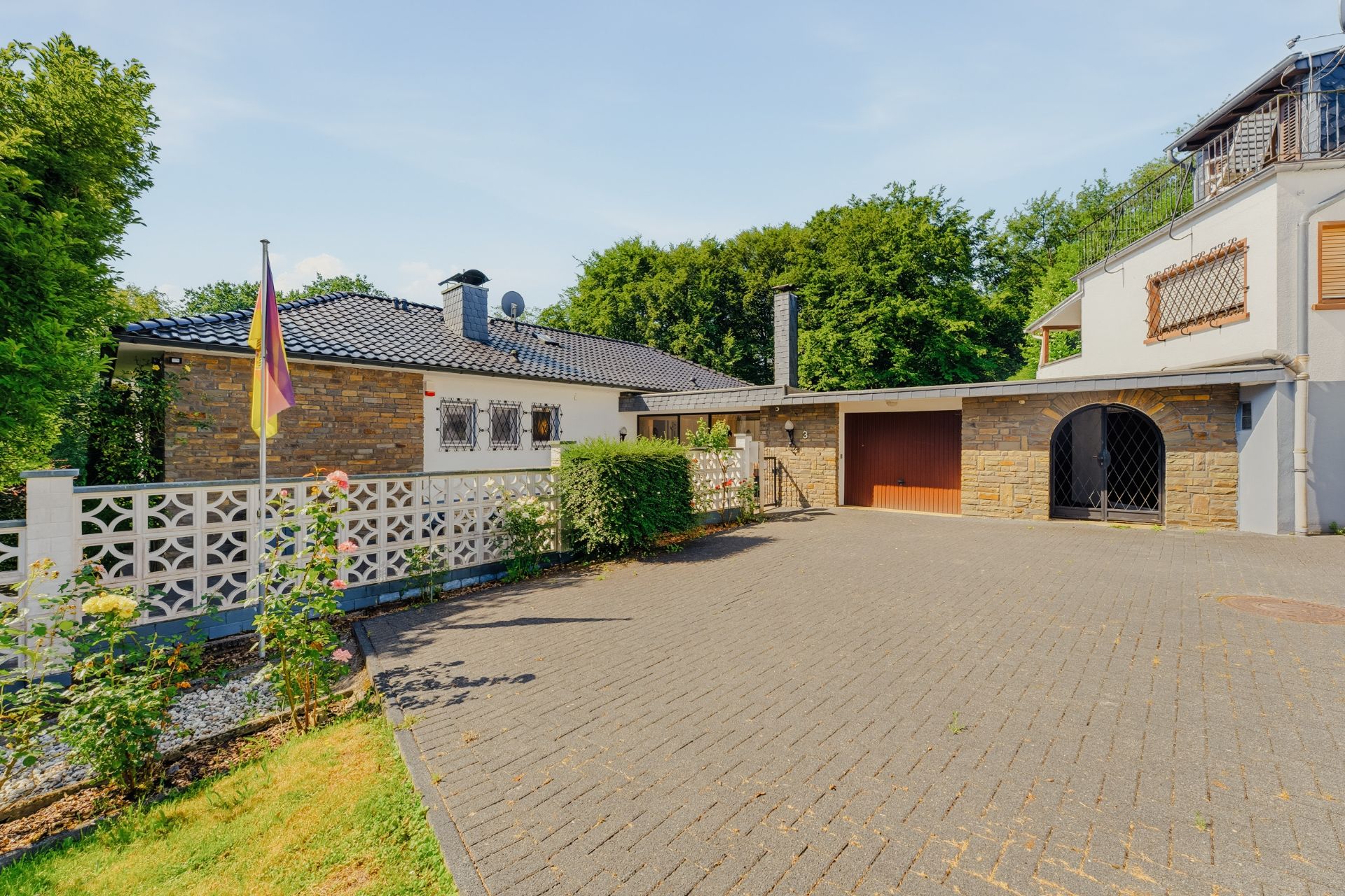 Gepflasterter Hof mit Garage, Wohnhaus und kleiner Garten mit Zaun, im Hintergrund Bäume.