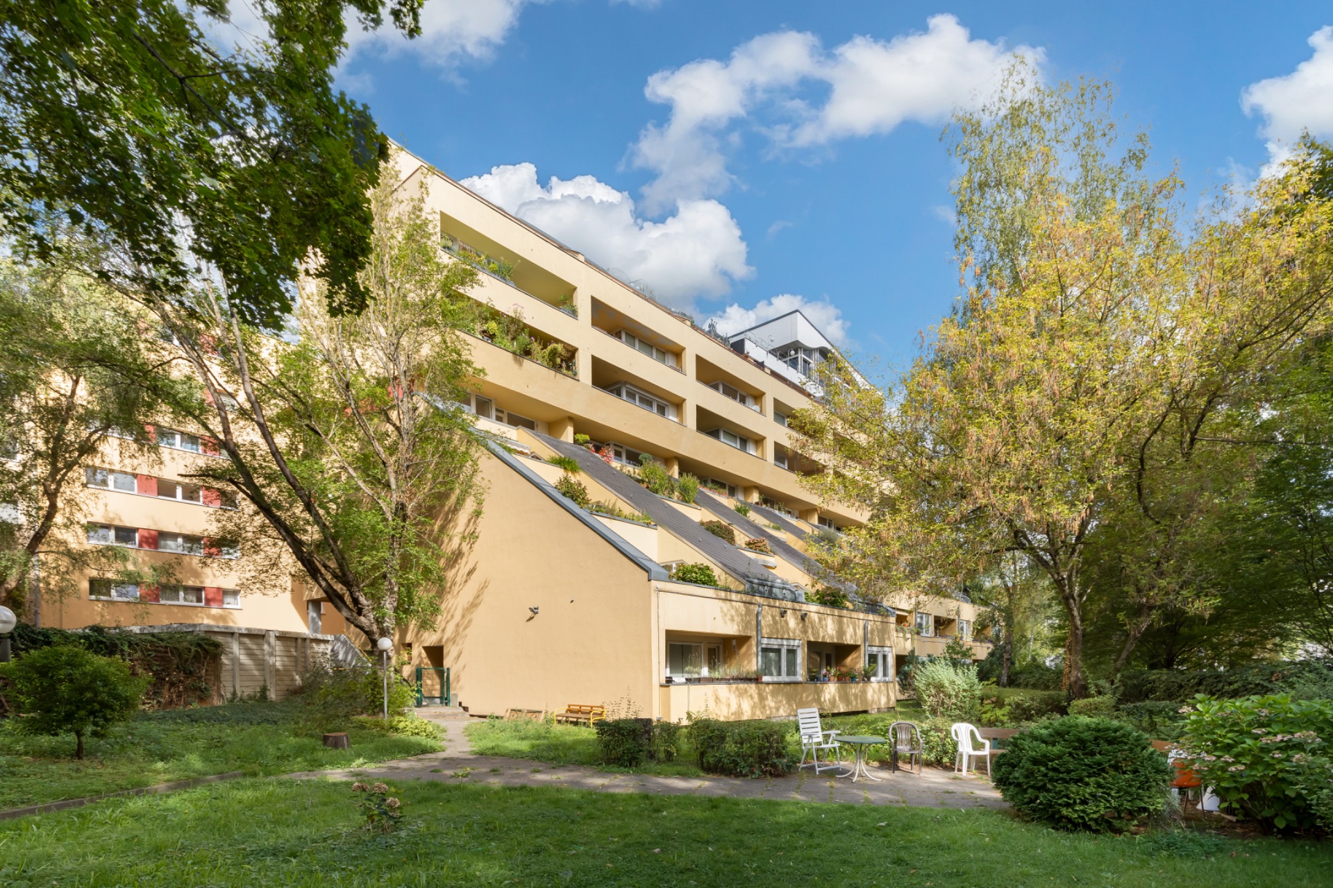 Mehrstöckiges Wohnhaus mit Balkonen und Gartenbereich, umgeben von Bäumen und Sitzmöglichkeiten.