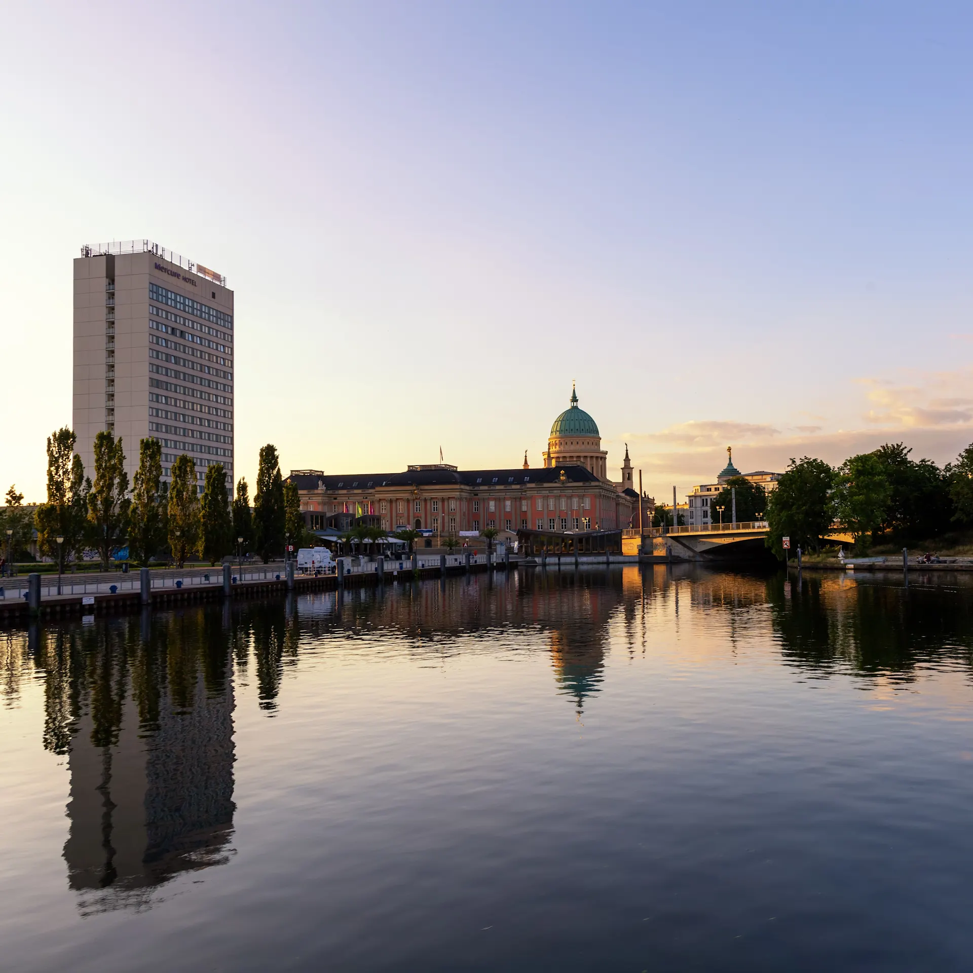 Hochhaus und Kuppelgebäude am Wasser bei Sonnenuntergang.