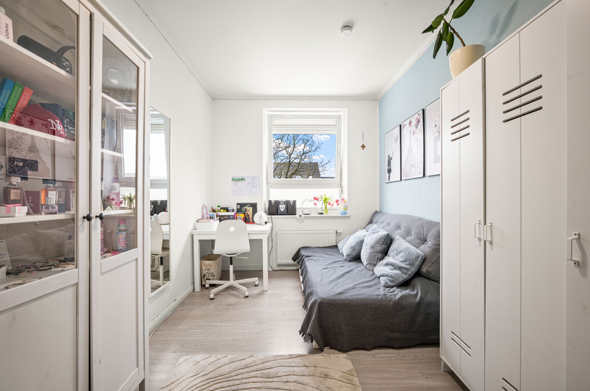 Helles Zimmer mit Sofa, Schreibtisch, Stuhl, Schrank und Regal, Pflanze auf dem Schrank.