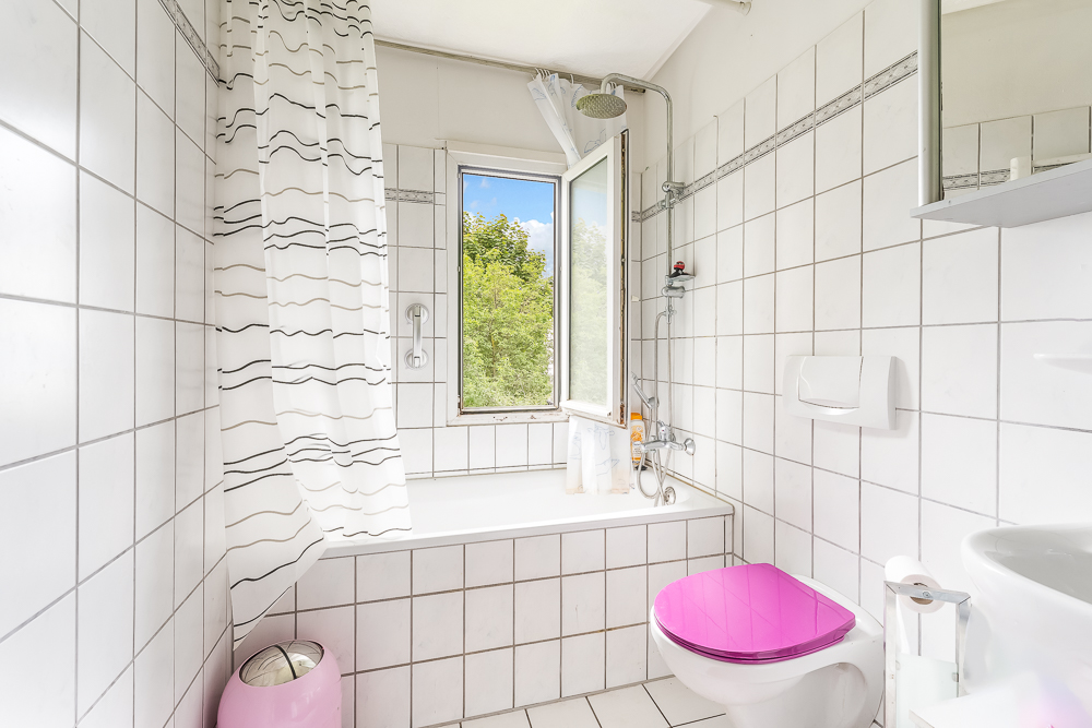 Helles Badezimmer mit Badewanne, Duschvorhang, offenem Fenster und rosa Toilettensitz.