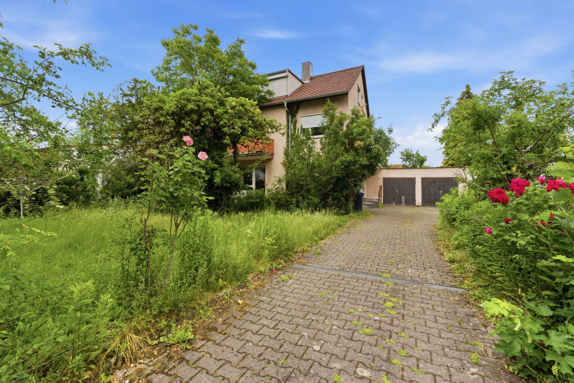 Gepflasterte Einfahrt zu einem Wohnhaus mit Garage, umgeben von grünem Garten und Blumen.