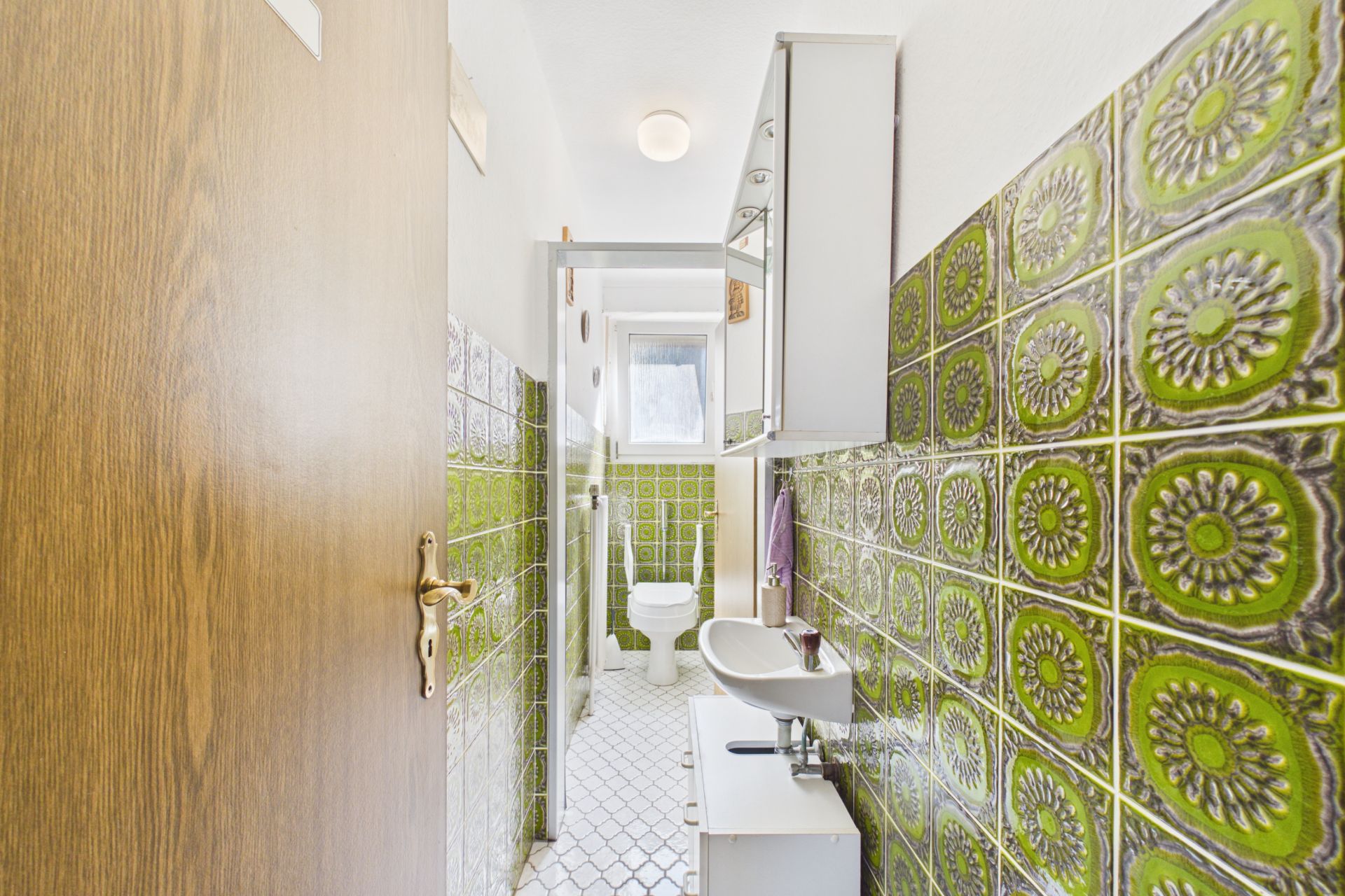 Schmales Bad mit grünen Fliesen, Waschbecken, Spiegel, Toilette und Fenster am Ende.