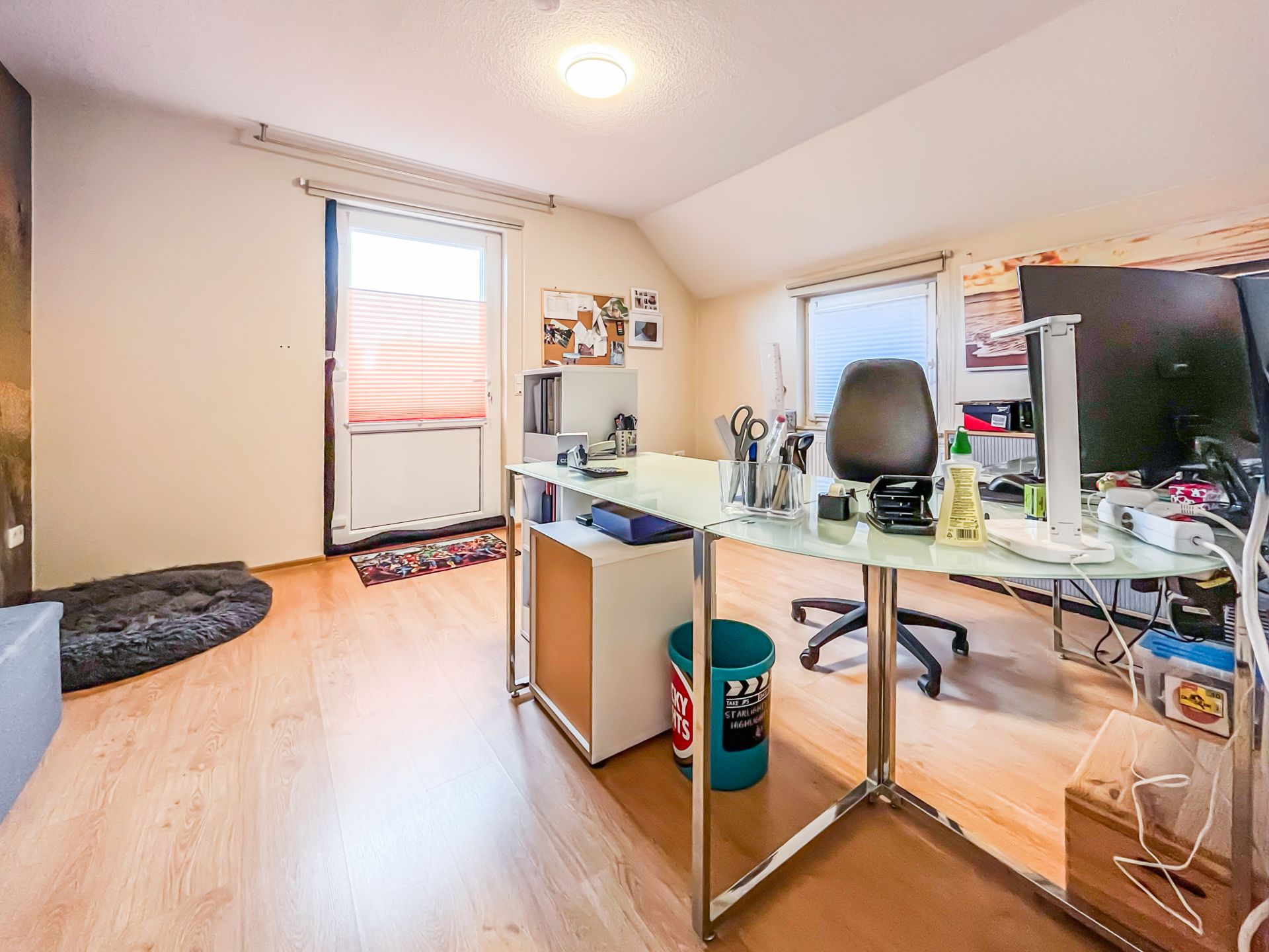 Helles Arbeitszimmer mit Schreibtisch, Stuhl, Computer, Fenster und Tür zu einem Balkon.
