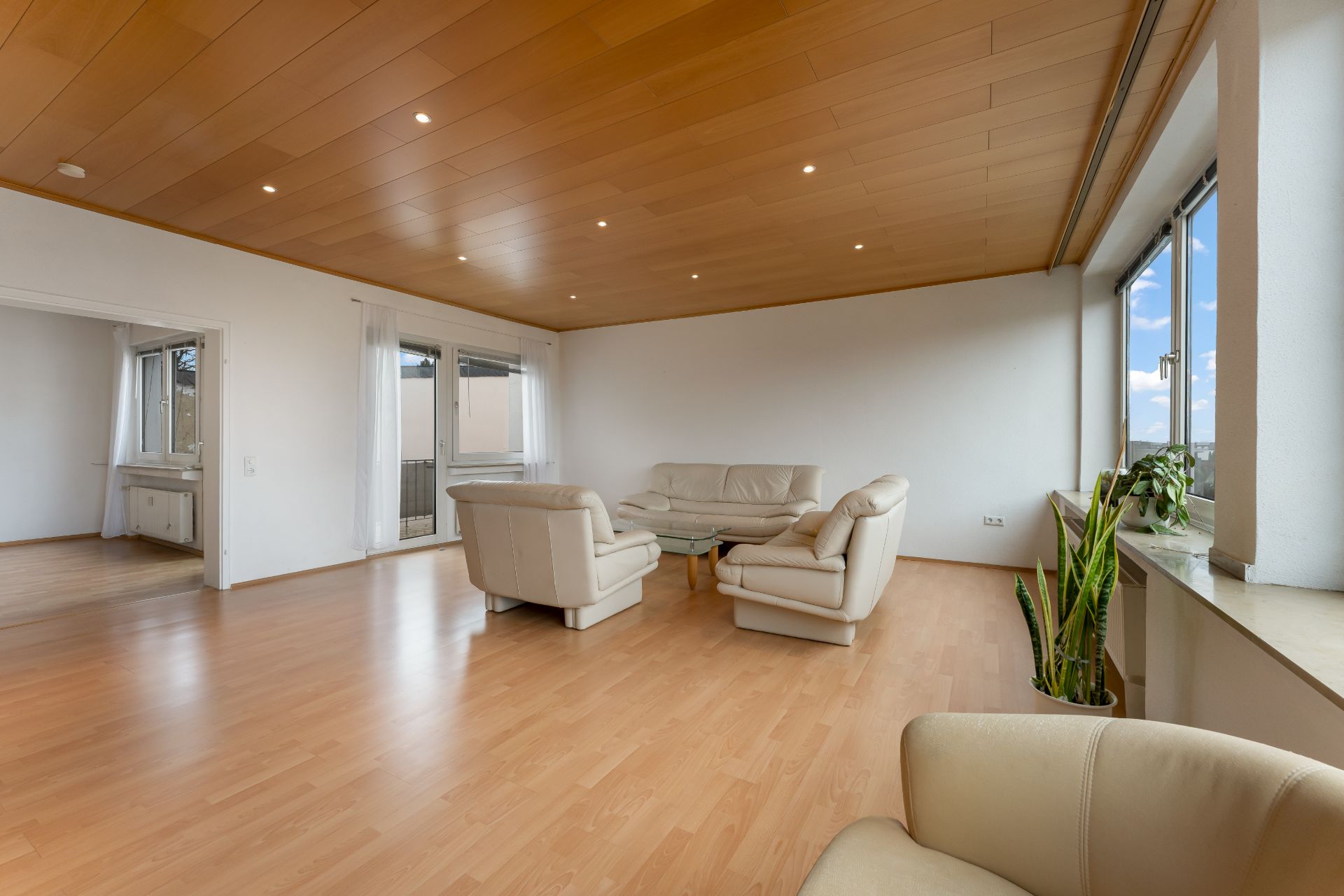 Helles Wohnzimmer mit hellen Ledersofas, Glastisch, Holzdecke, großen Fenstern und Pflanzen.
