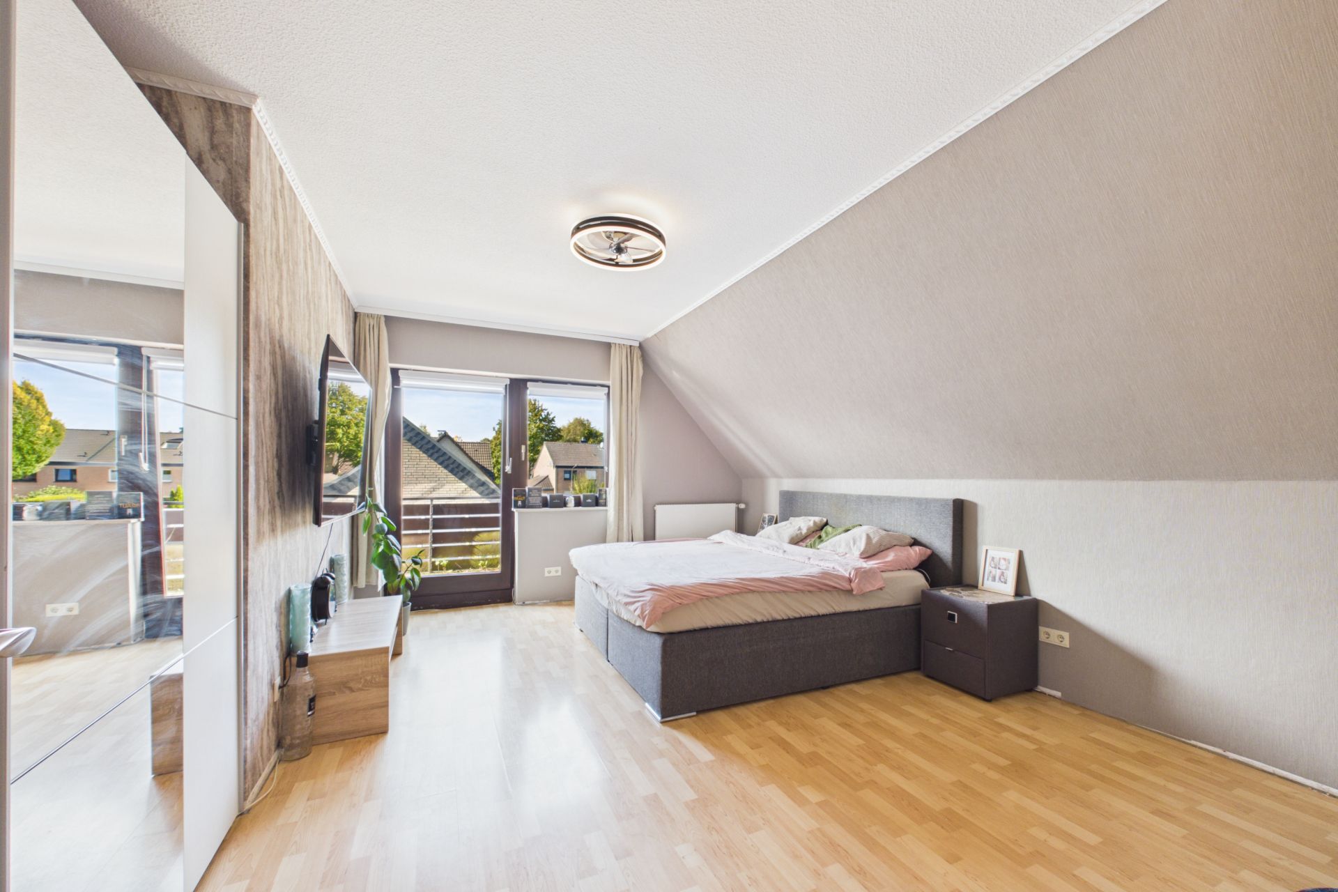 Helles Schlafzimmer mit Doppelbett, Fernseher, großem Fenster und Holzfußboden.