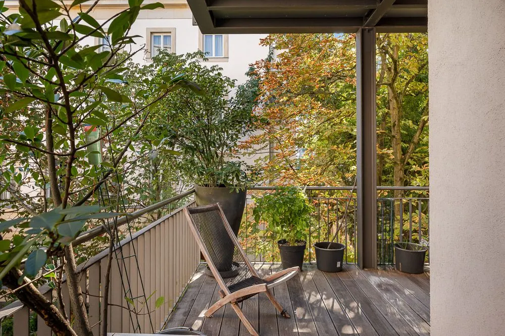Holzbalkon mit Stuhl und mehreren Pflanzen in Töpfen, Blick auf Bäume und Nachbargebäude.