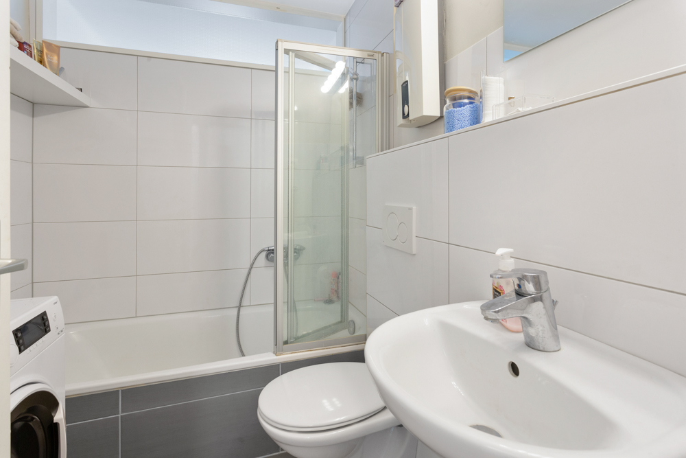 Modernes Badezimmer mit Dusche, Waschbecken, Waschmaschine und weißen Fliesen.