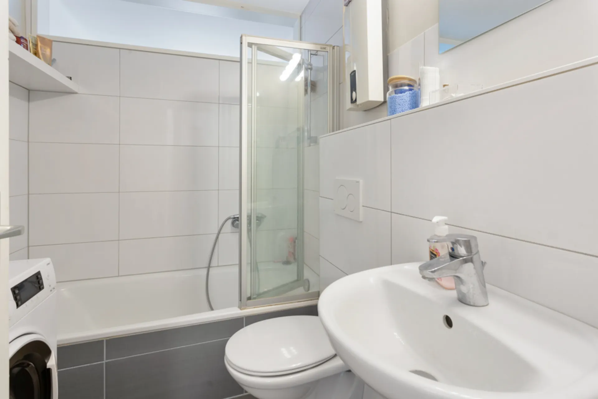 Modernes Badezimmer mit Dusche, Waschbecken, Waschmaschine und weißen Fliesen.