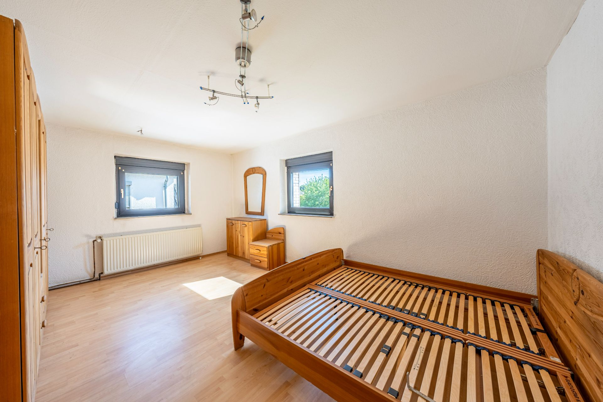 Helles Schlafzimmer mit Holzkleiderschrank, Lattenrostbett und Kommode mit Spiegel.