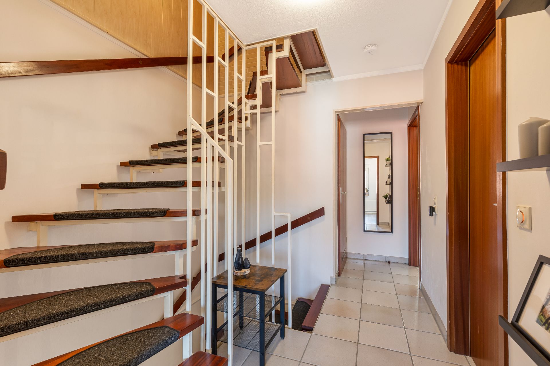 Flur mit offener Holztreppe, weißer Wand, Spiegel und Türrahmen aus Holz.