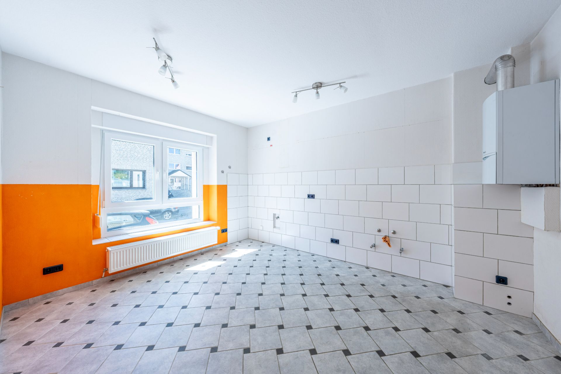 Leerer, gefliester Raum mit weiß-orangen Wänden, großem Fenster und Heizkörper.