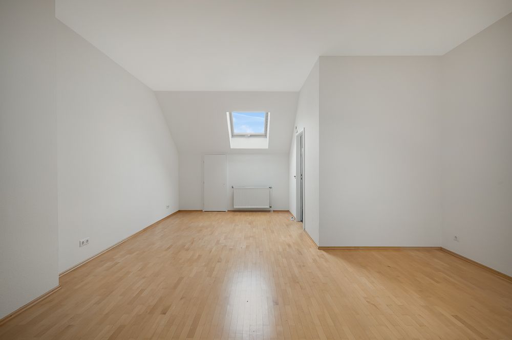 Leerer Raum mit hellem Holzboden, weißem Wänden und Dachfenster.