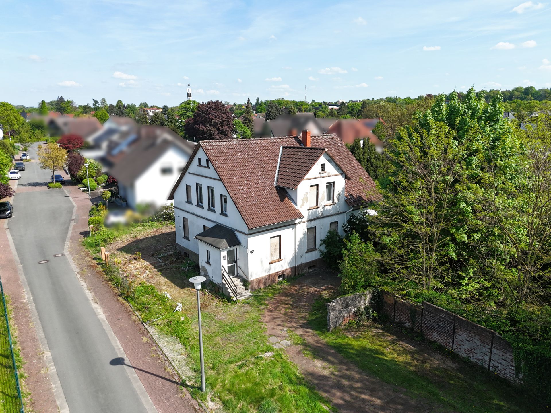 Weißes Einfamilienhaus mit rotem Dach an einer ruhigen Straße, umgeben von Bäumen und Grünfläche.
