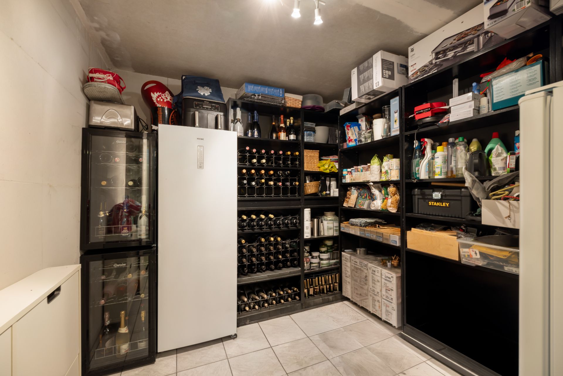 Lagerraum mit Kühlschrank, Weinregalen und schwarzen Regalen voller Vorräte und Haushaltswaren.
