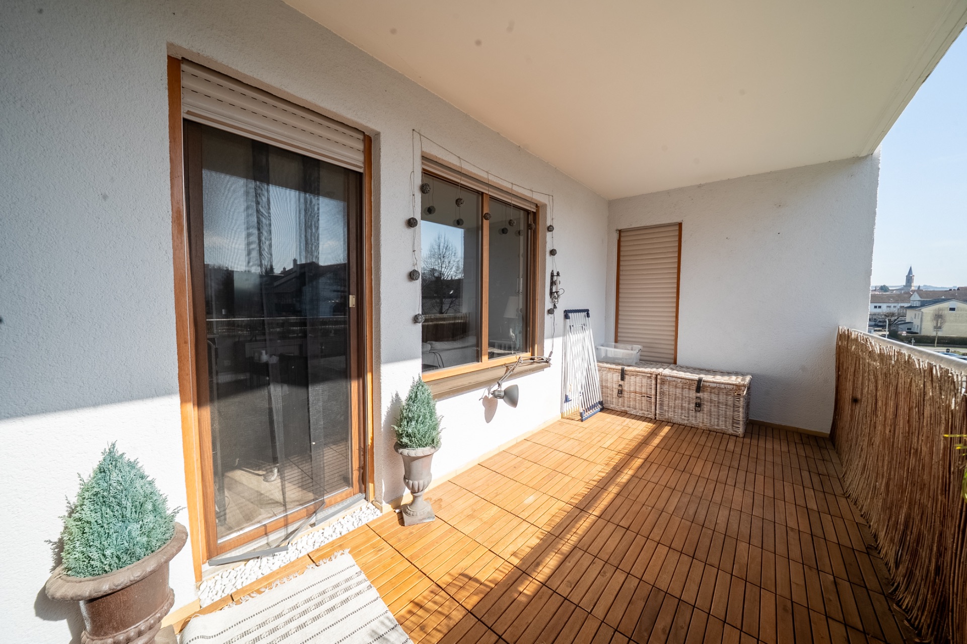 Überdachter Balkon mit Holzboden, zwei Pflanzen, Truhe und Blick auf Nachbargebäude.