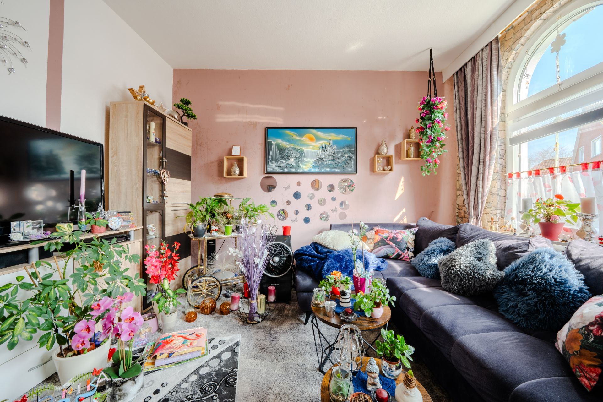 Bunt dekoriertes Wohnzimmer mit vielen Pflanzen, großem Sofa und rundem Fenster.