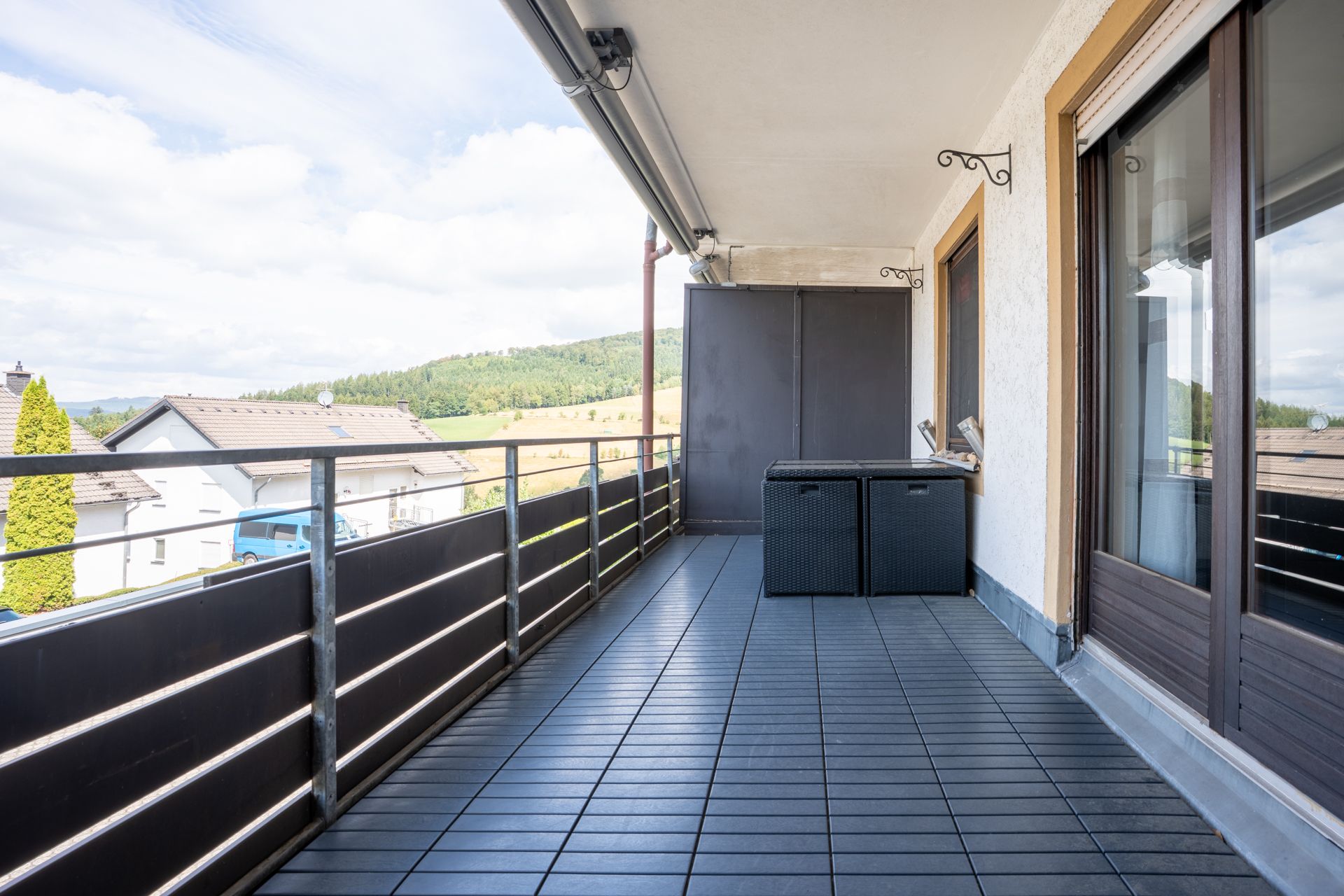 Großer Balkon mit Holzboden, Geländer und zwei dunklen Sitztruhen, Blick auf Häuser und grüne Hügel.