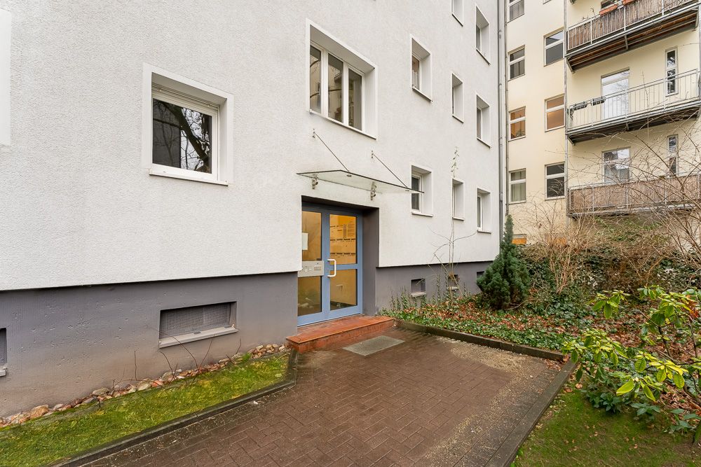 Eingang eines Wohnhauses mit Glasdach, umgeben von Gehweg und Pflanzen.