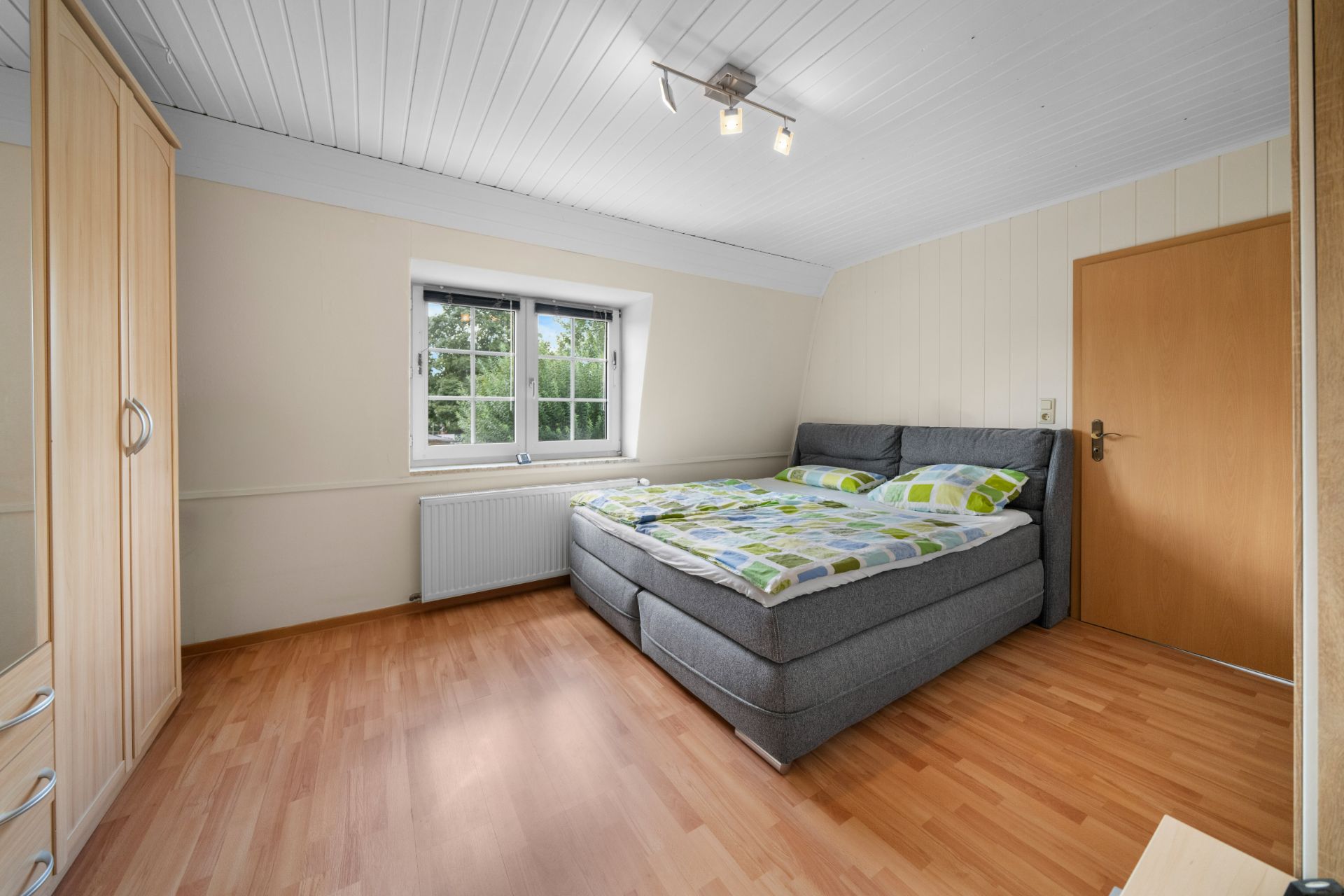 Helles Schlafzimmer mit Doppelbett, Holzschrank, Fenster und Holzboden.