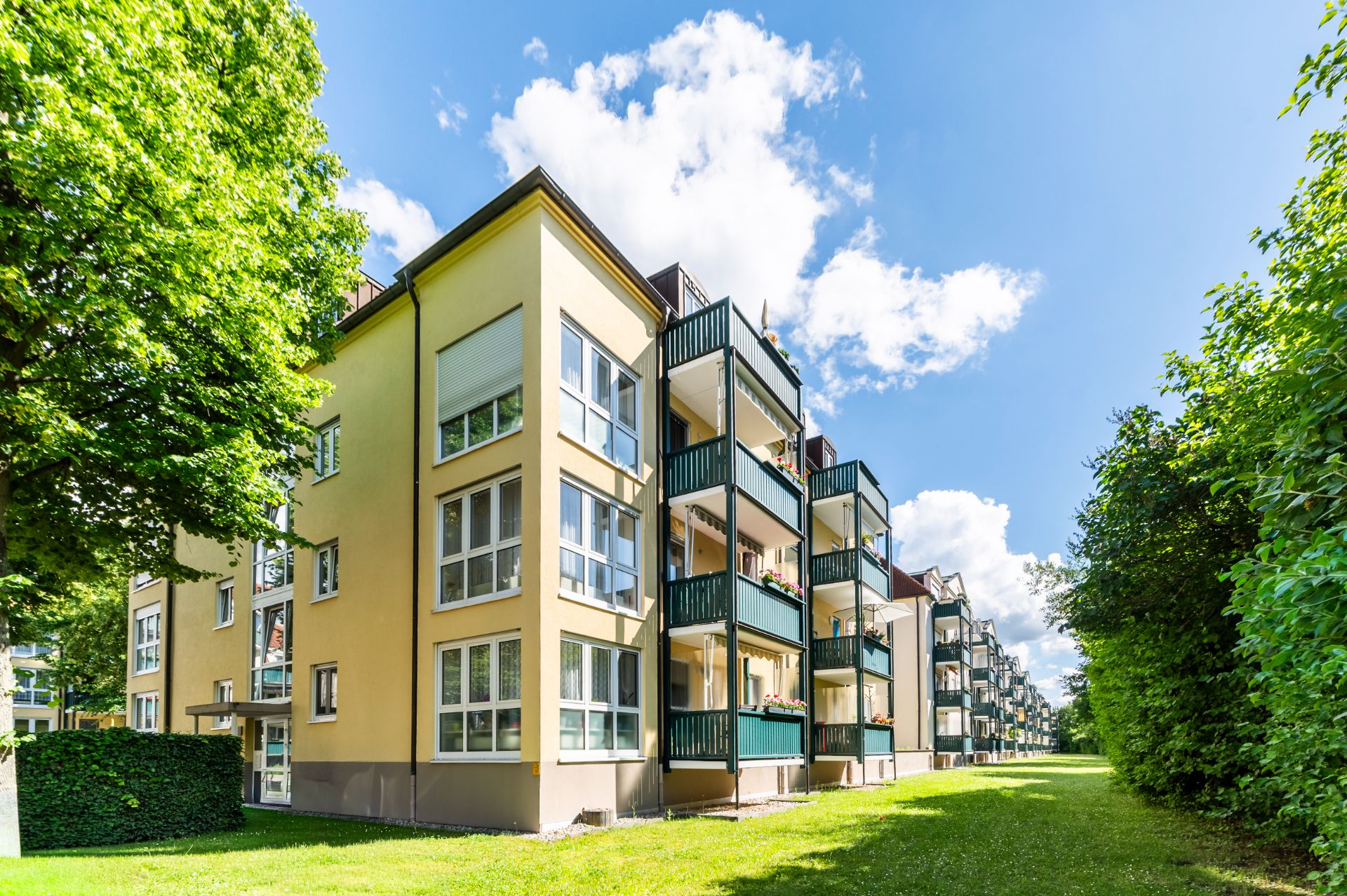Mehrfamilienhaus mit mehreren Balkonen, davor eine große Wiese und Bäume.