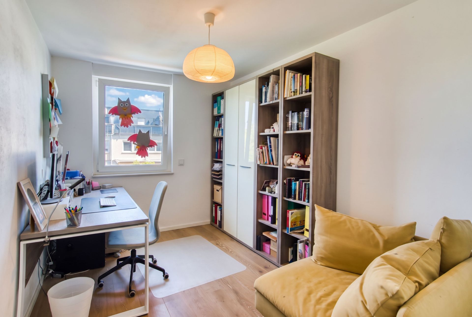 Helles Arbeitszimmer mit Schreibtisch, Bücherregal, gelbem Sessel und Fenster mit Eulen-Dekoration.