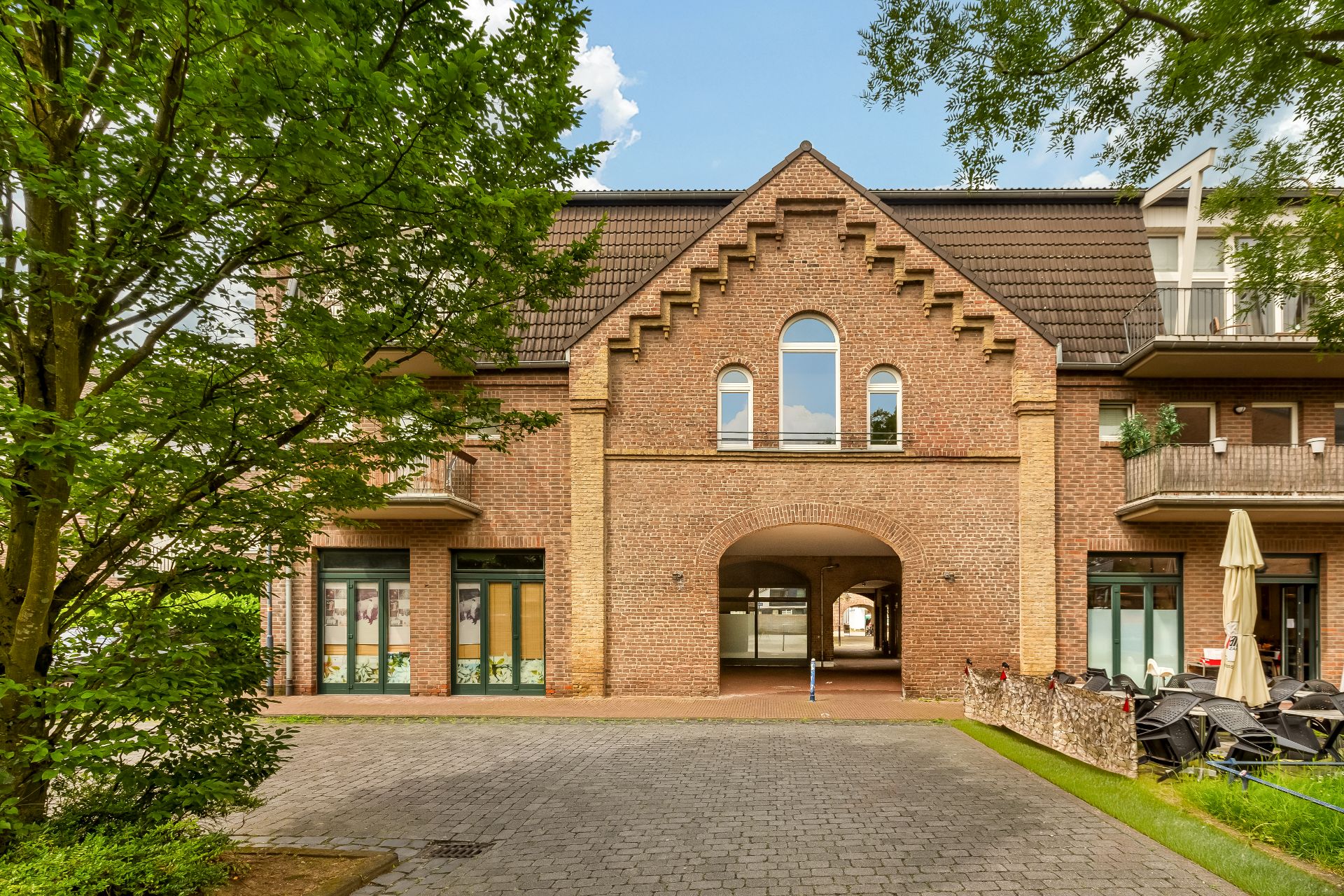 Backsteinwohnhaus mit Rundbogenpassage, Fenstern und Balkonen, im Vordergrund Bäume und Terrasse.