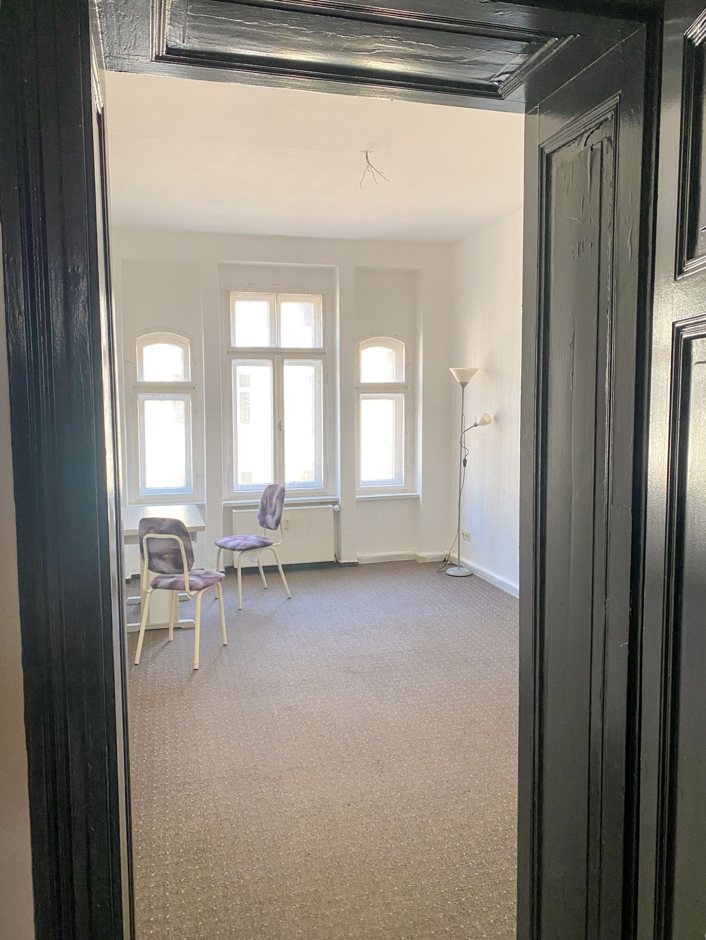 Helles Zimmer mit drei Fenstern, zwei Stühlen, kleinem Tisch und Stehlampe, durch eine Tür fotografiert.
