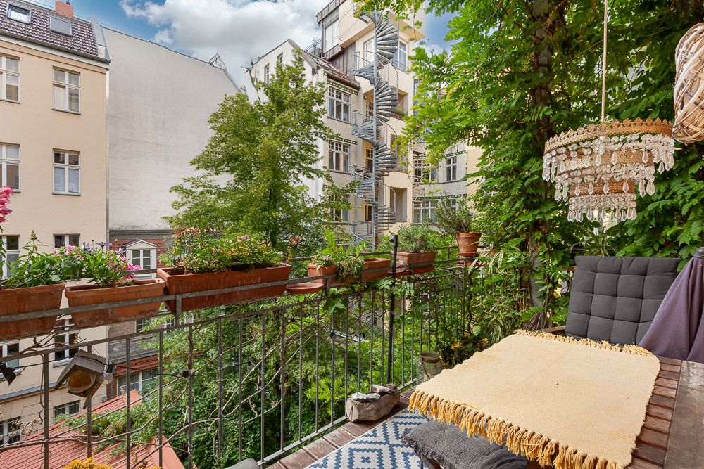 Balkon mit Pflanzen, einer Sitzgelegenheit, Teppich und Hängelampe, Blick auf umliegende Wohnhäuser.