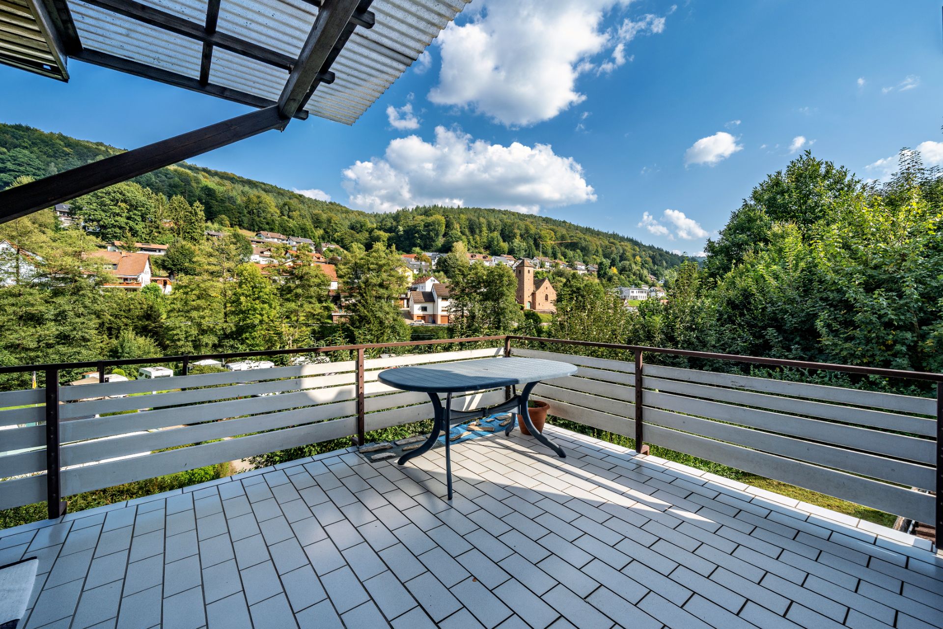 Große Terrasse mit Tisch, Ausblick auf grüne Hügel und Wohnhäuser im Hintergrund.