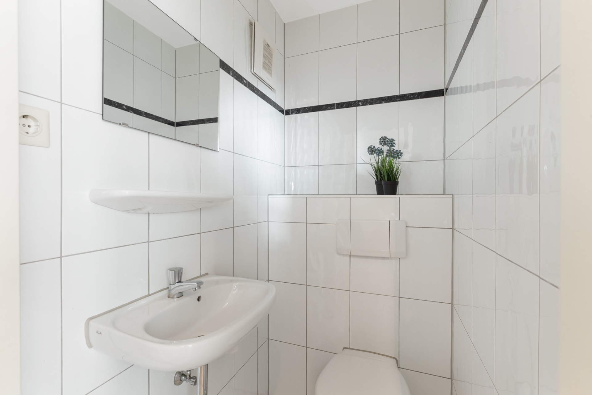 Kleines, weiß gefliestes Badezimmer mit Waschbecken, Spiegel, WC und einer Pflanze auf der Ablage.