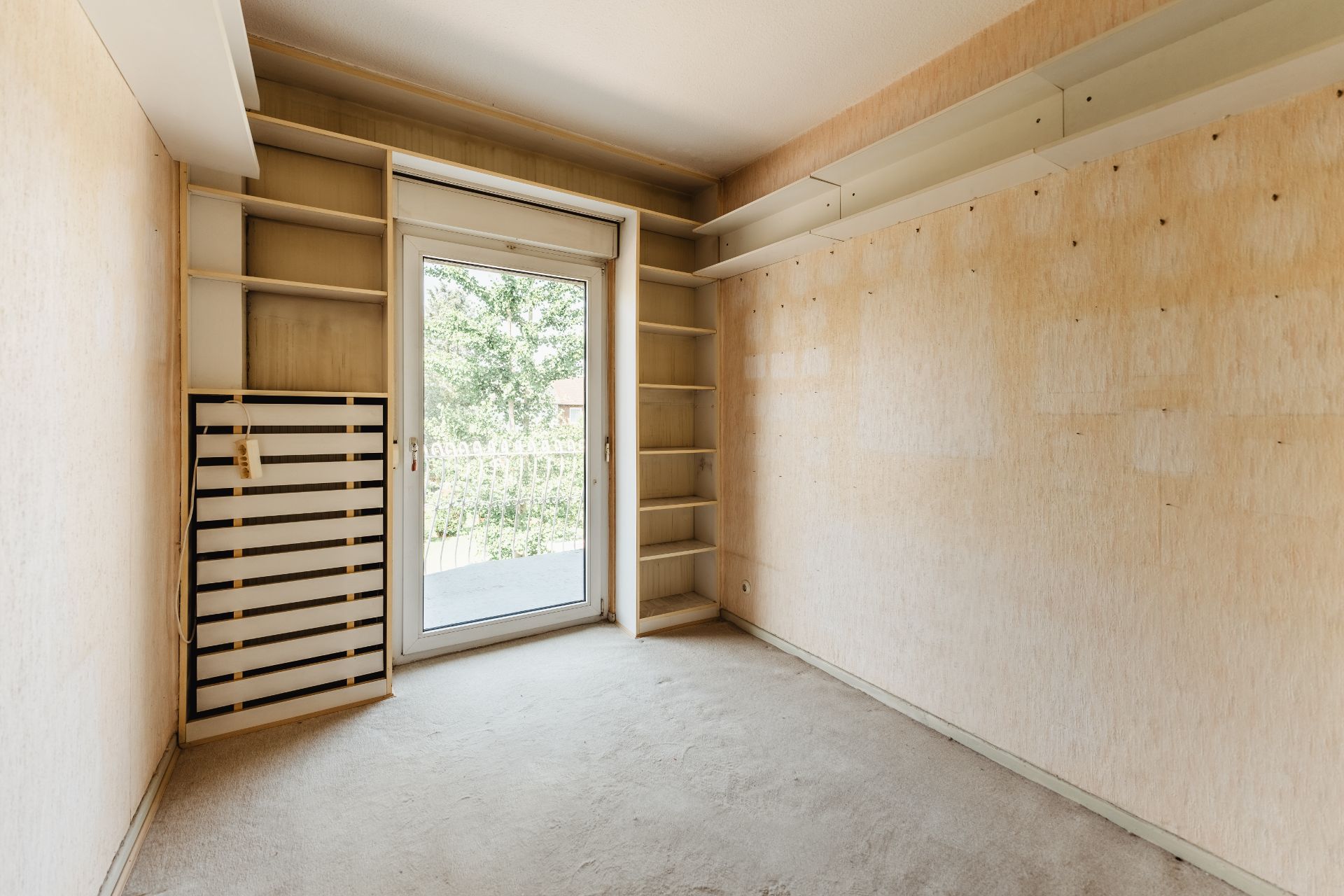 Leeres Zimmer mit Regal und Tür zum Balkon, helle Wände und Teppichboden.