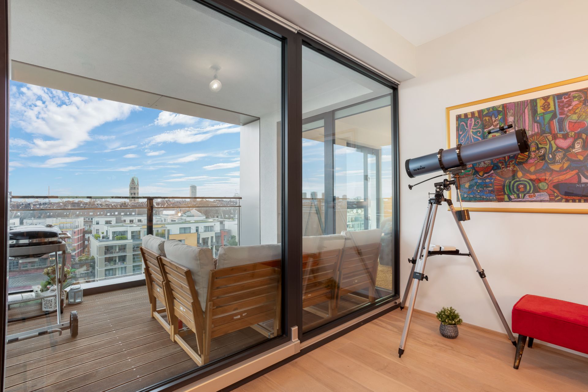 Wohnzimmer mit Teleskop vor Fenster, dahinter Balkon mit Stühlen und Stadtblick.