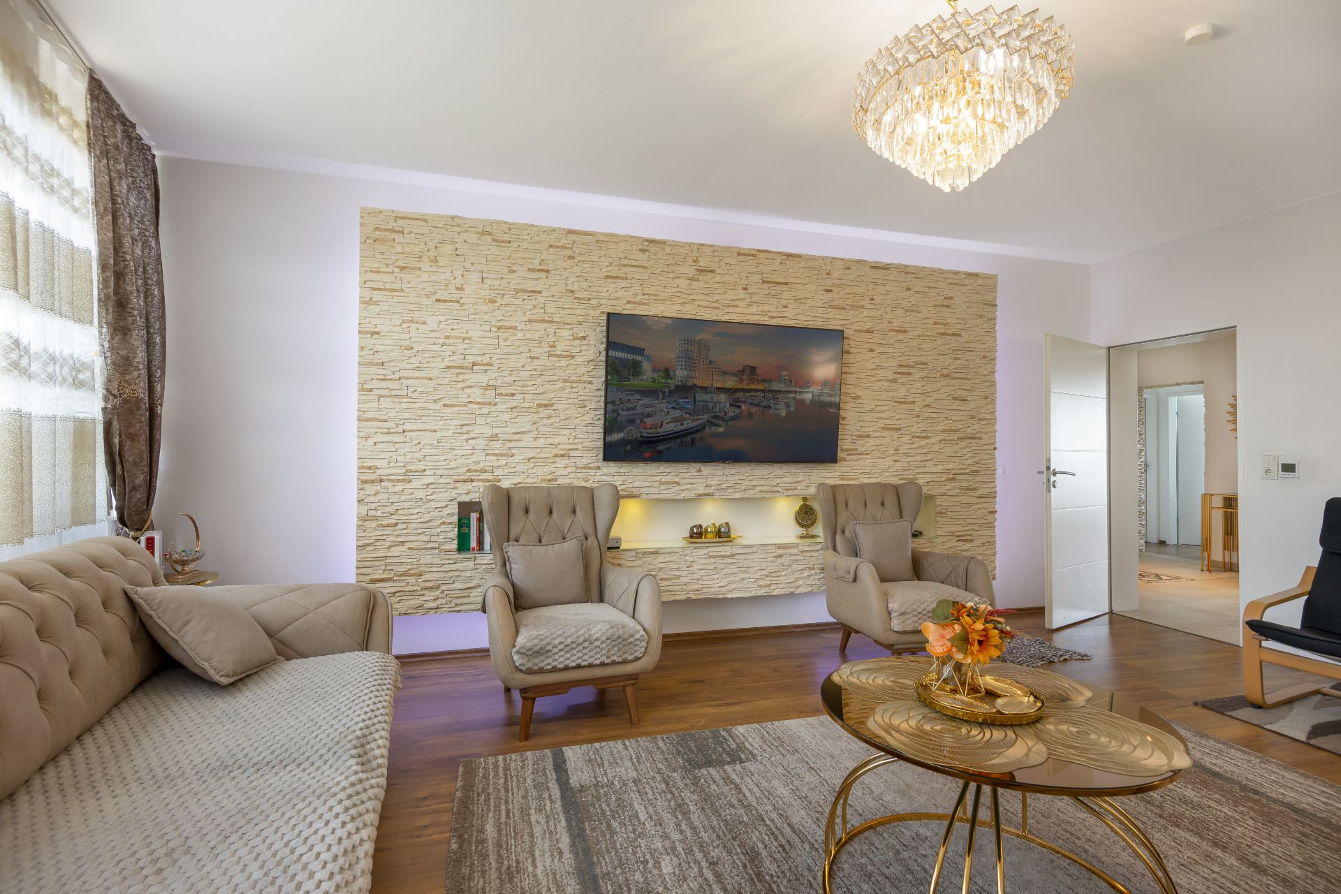 Helles Wohnzimmer mit Sofa, Sesseln, Couchtisch, Teppich und Fernseher an einer Steinwand.