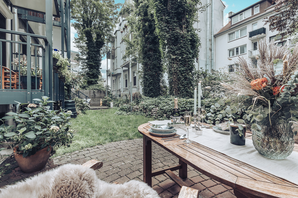 Gedeckter Holztisch im Garten mit Blumen, Geschirr und Kerzen, umgeben von Wohnhäusern und viel Grün.