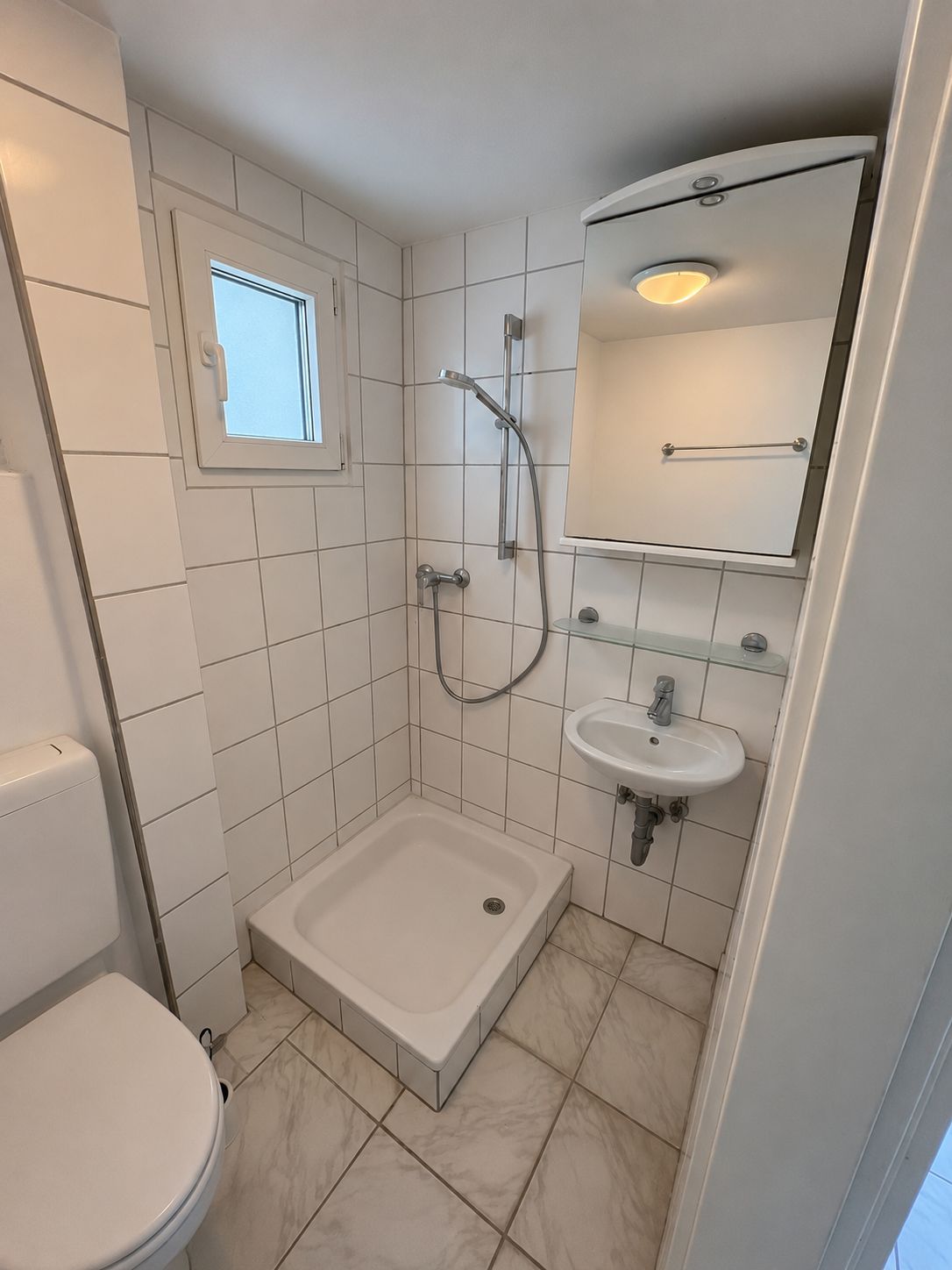 Kleines Badezimmer mit Dusche, kleinem Waschbecken, Spiegel, Toilette und Fenster.