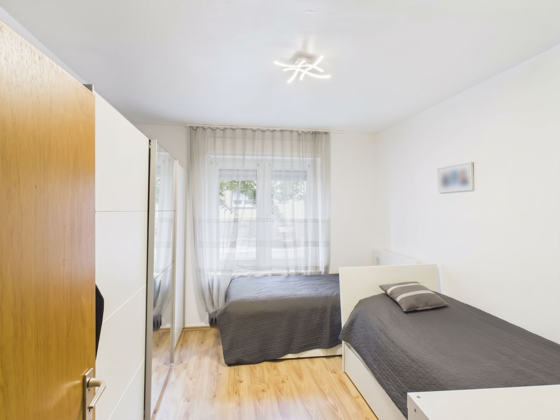 Helles Schlafzimmer mit zwei Einzelbetten, einem Schrank und einem Fenster mit Gardine.