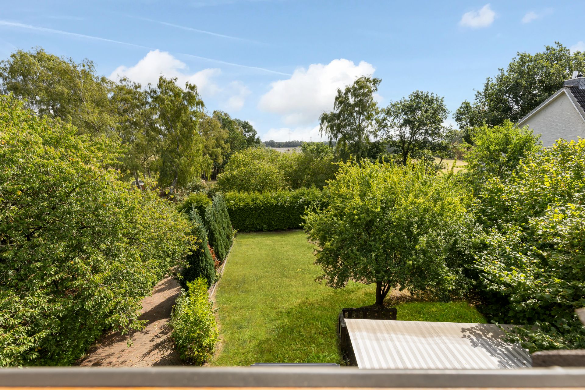Blick auf einen grünen Garten mit Bäumen und Rasen, im Hintergrund sind Felder und Bäume sichtbar.