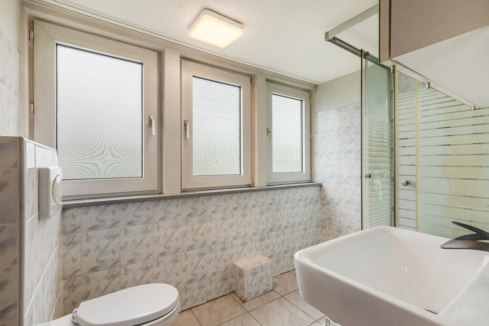 Helles Badezimmer mit drei Fenstern, Toilette, Waschbecken und Dusche mit Glaswand.
