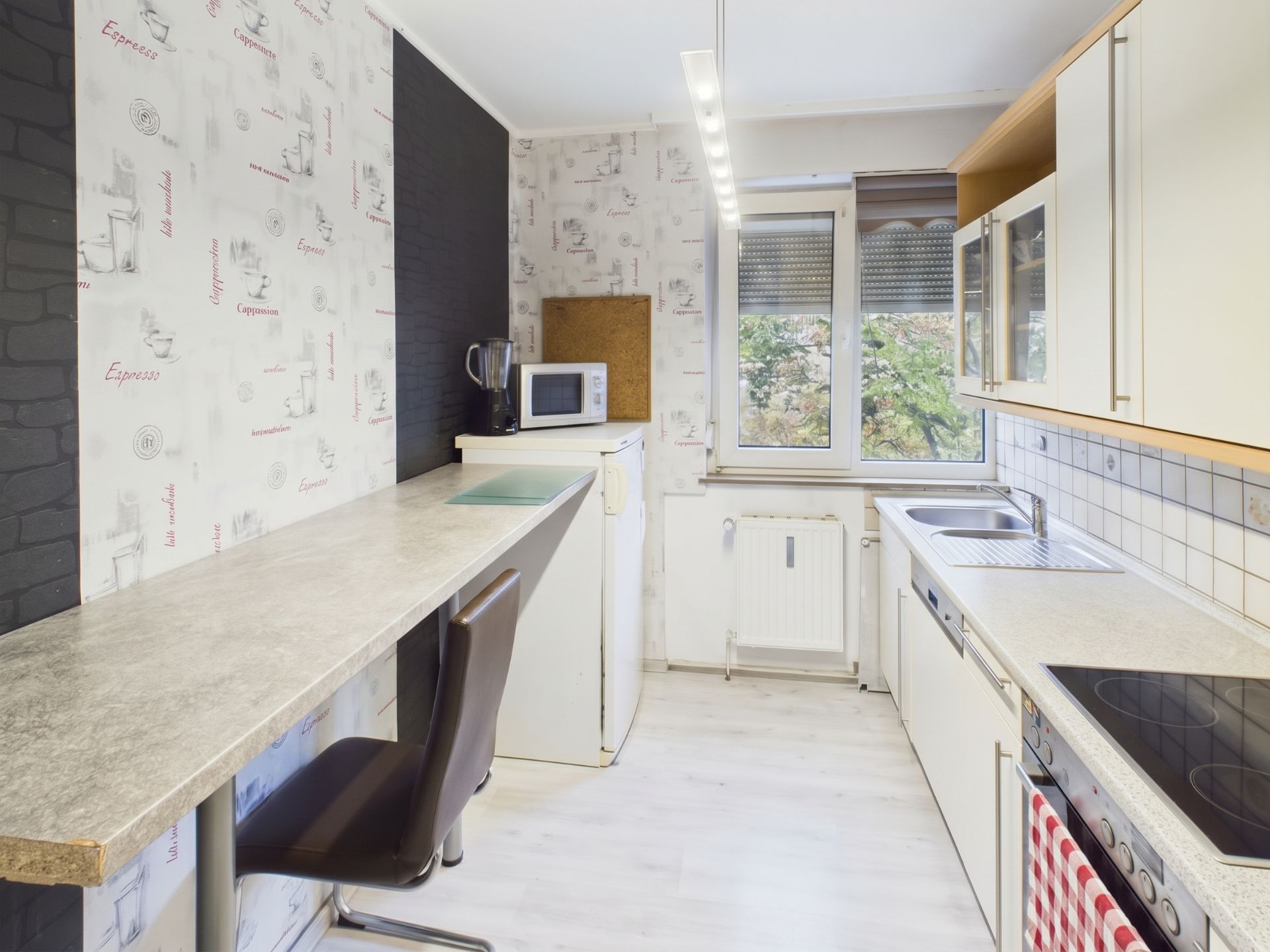 Helle Küche mit langer Arbeitsplatte, Stühlen, Herd, Spüle, Kühlschrank und Fenster.