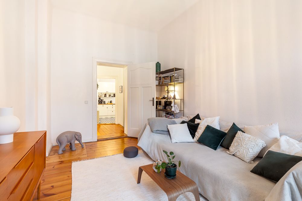 Wohnzimmer mit Sofa, vielen Kissen, Teppich, Holztisch und Regal. Tür führt in einen weiteren Raum.