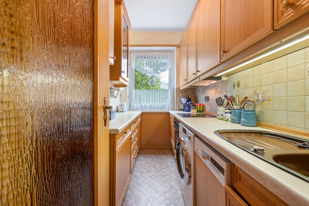 Schmale Küche mit Holzschränken, Waschmaschine, Spüle, Fenster und Geräten auf der Arbeitsplatte.