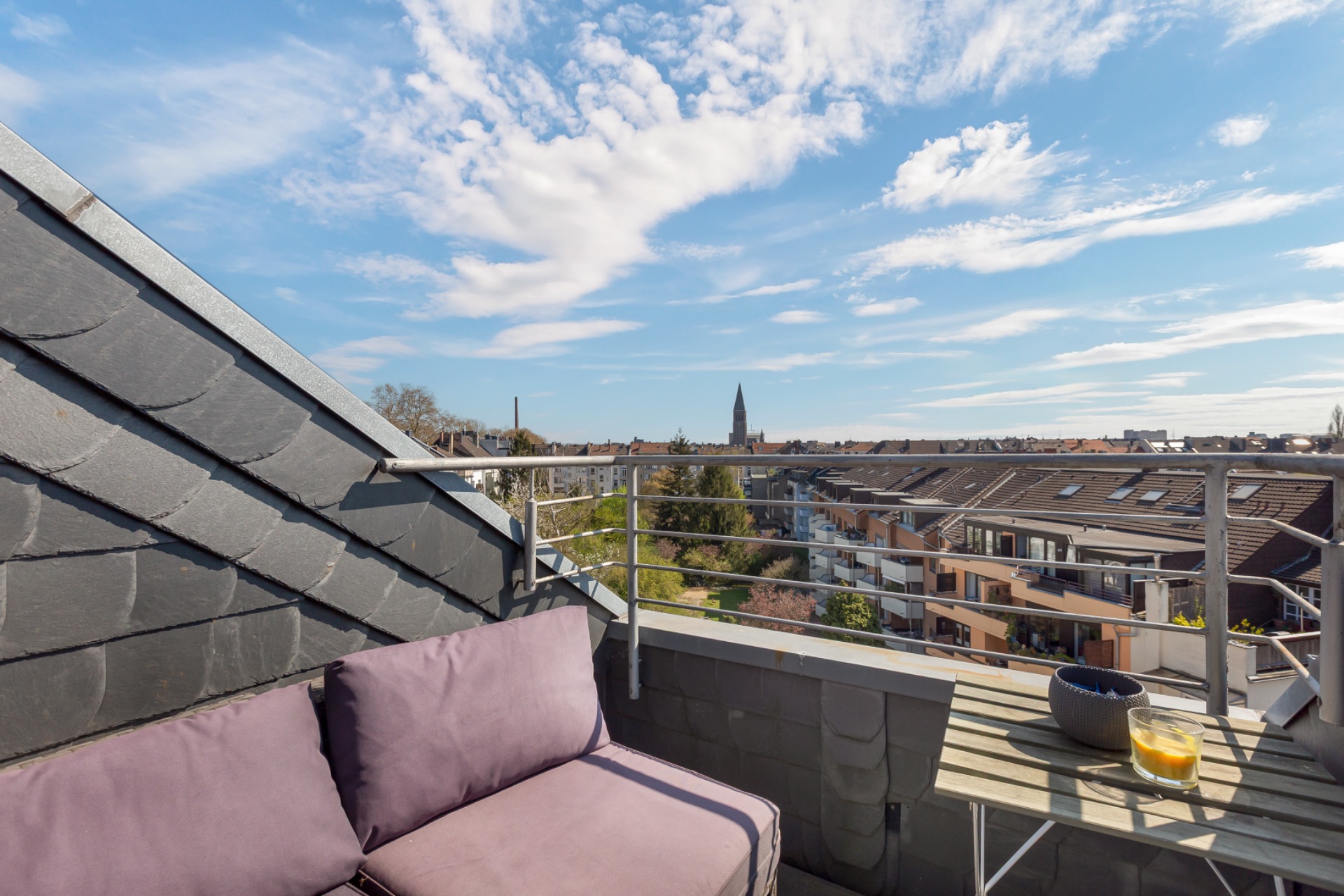 Kleine Dachterrasse mit lila Sofa, Tisch, Kerze und Stadtblick bei blauem Himmel.
