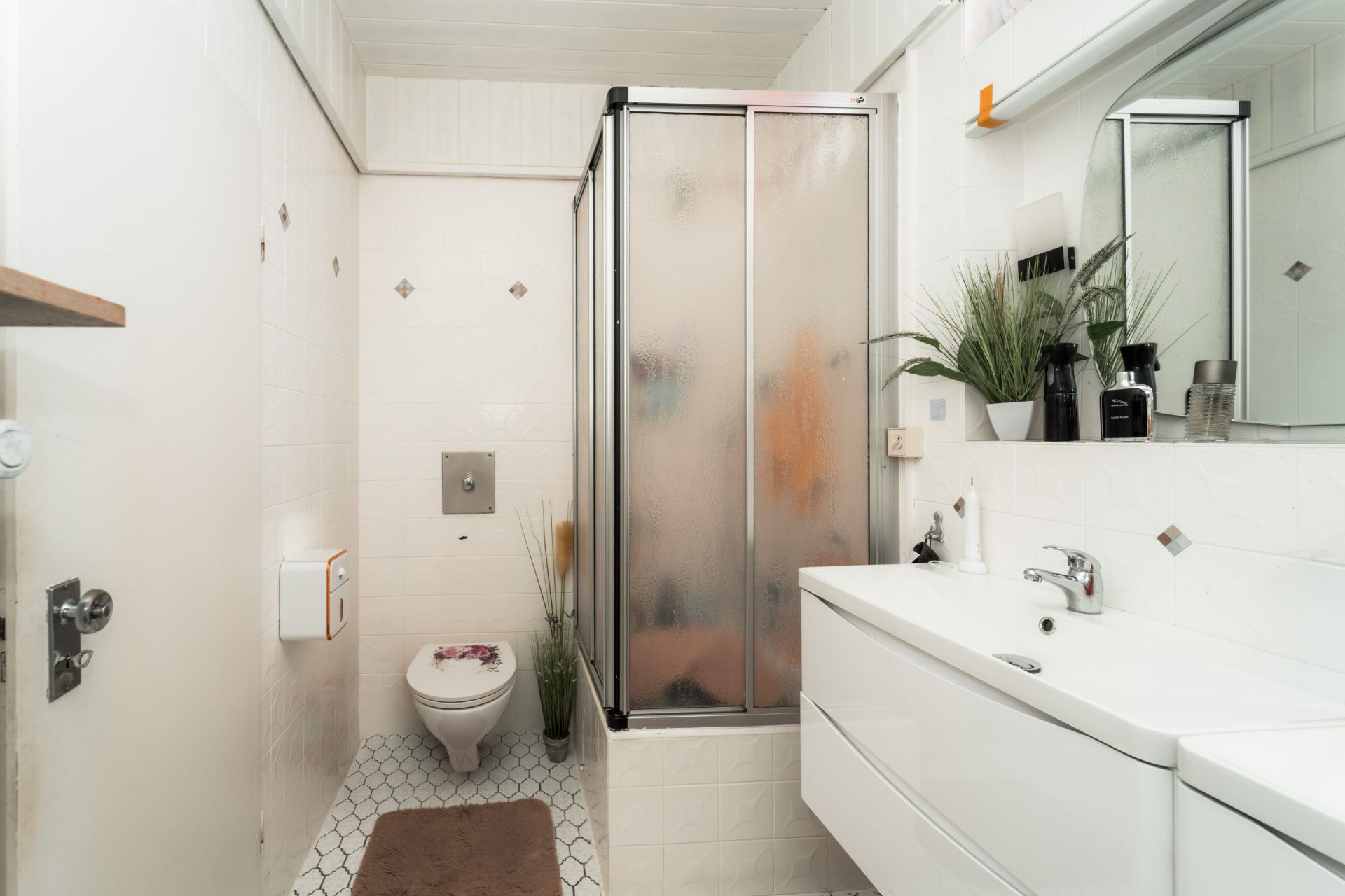 Helles Badezimmer mit Dusche, WC, Waschbecken und Spiegel. Pflanzen und Dekorationen sind sichtbar.