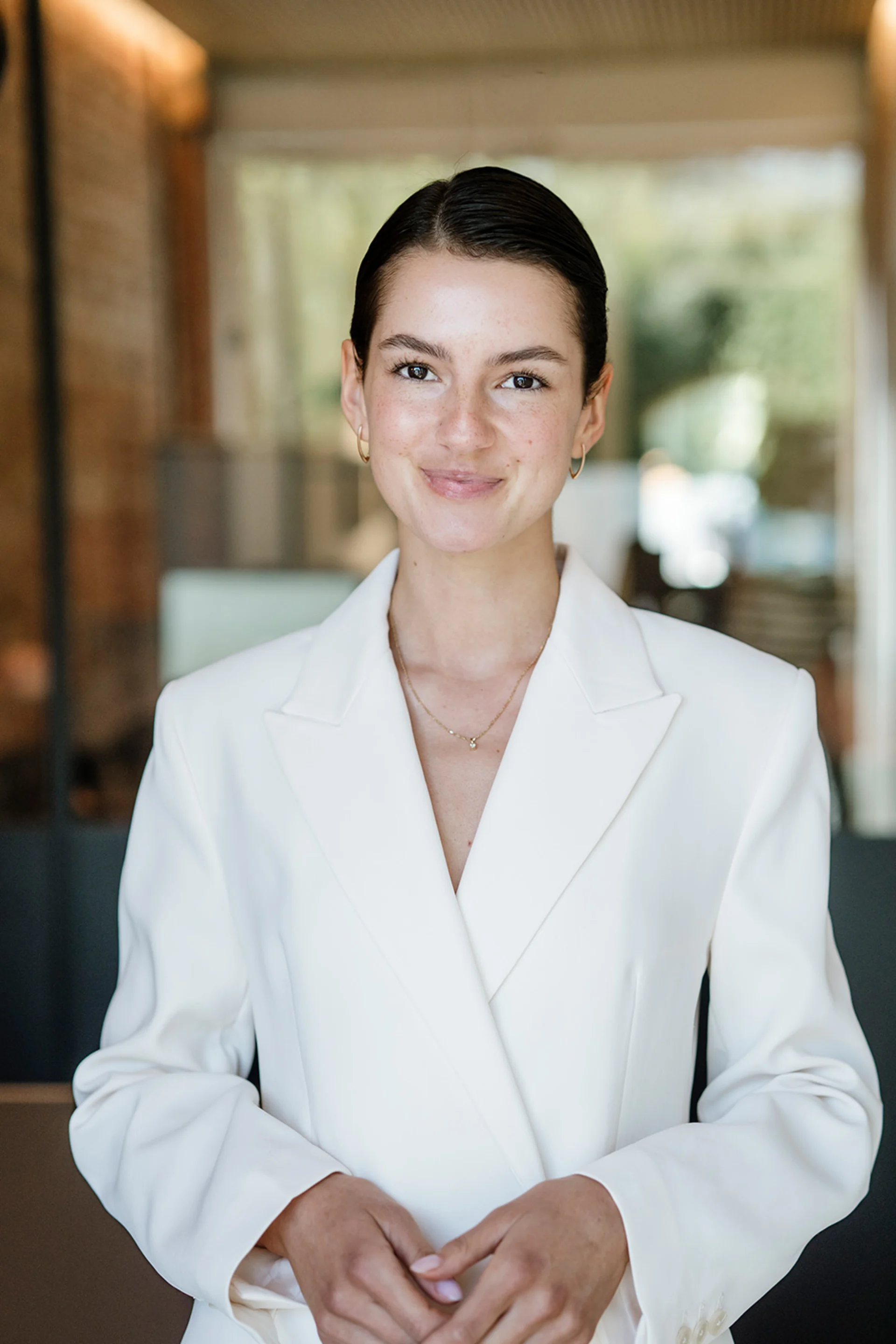 Junge Frau mit weißem Blazer steht in einem modernen Raum mit großen Fenstern.