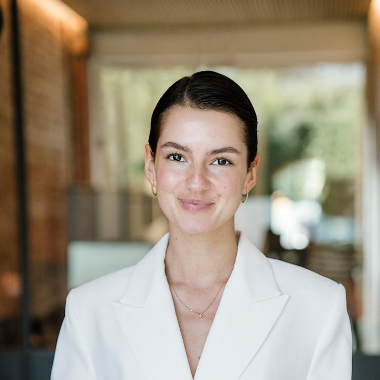 Junge Frau mit weißem Blazer steht in einem modernen Raum mit großen Fenstern.