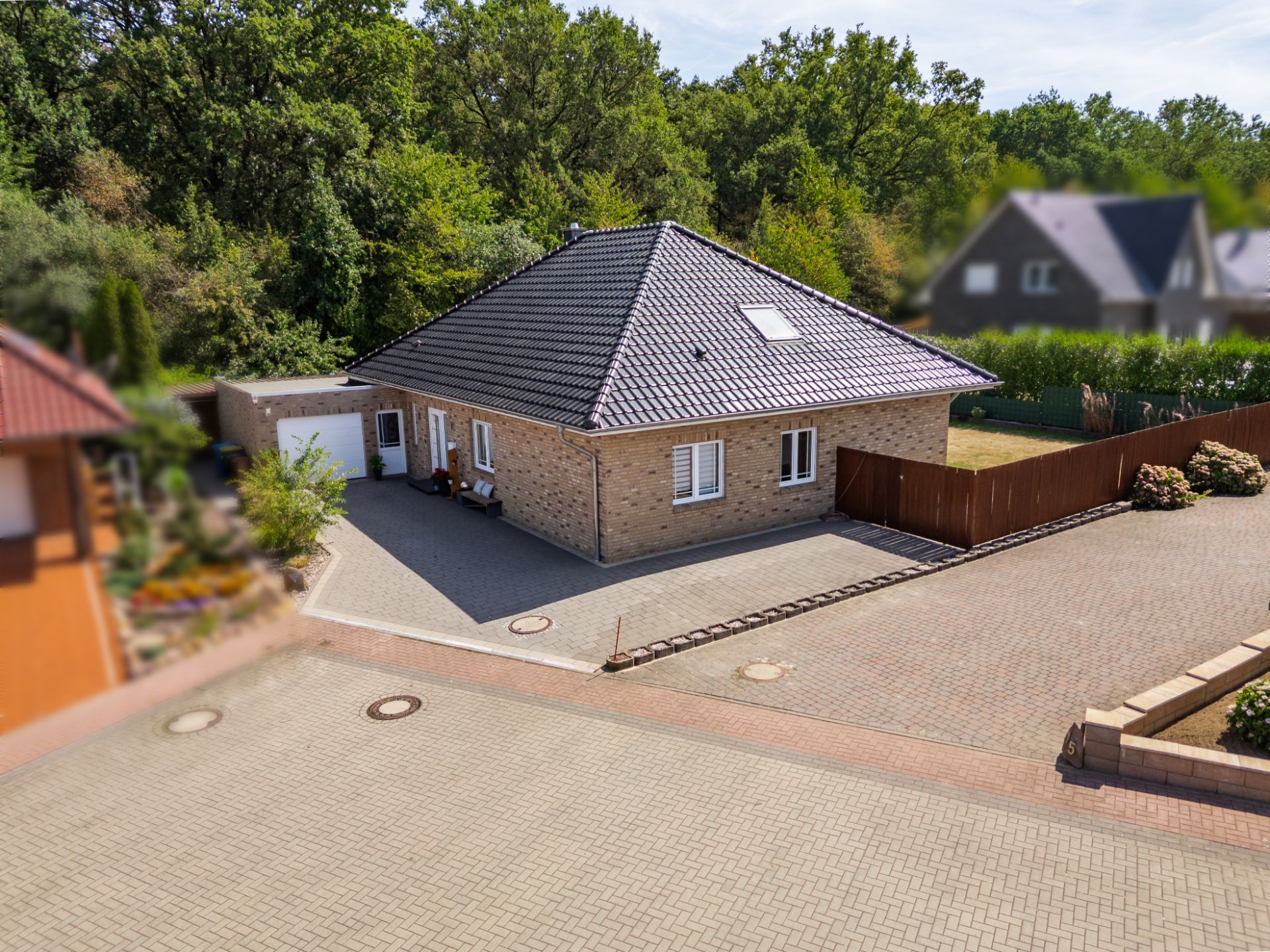 Einfamilienhaus mit Ziegeldach und gepflastertem Hof, umgeben von Bäumen und Nachbarhäusern.