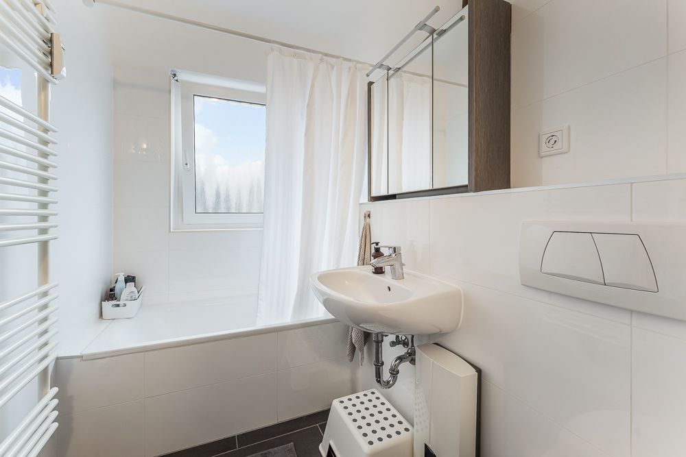 Helles Bad mit Waschbecken, Spiegel, Badewanne, Fenster und weißem Duschvorhang.